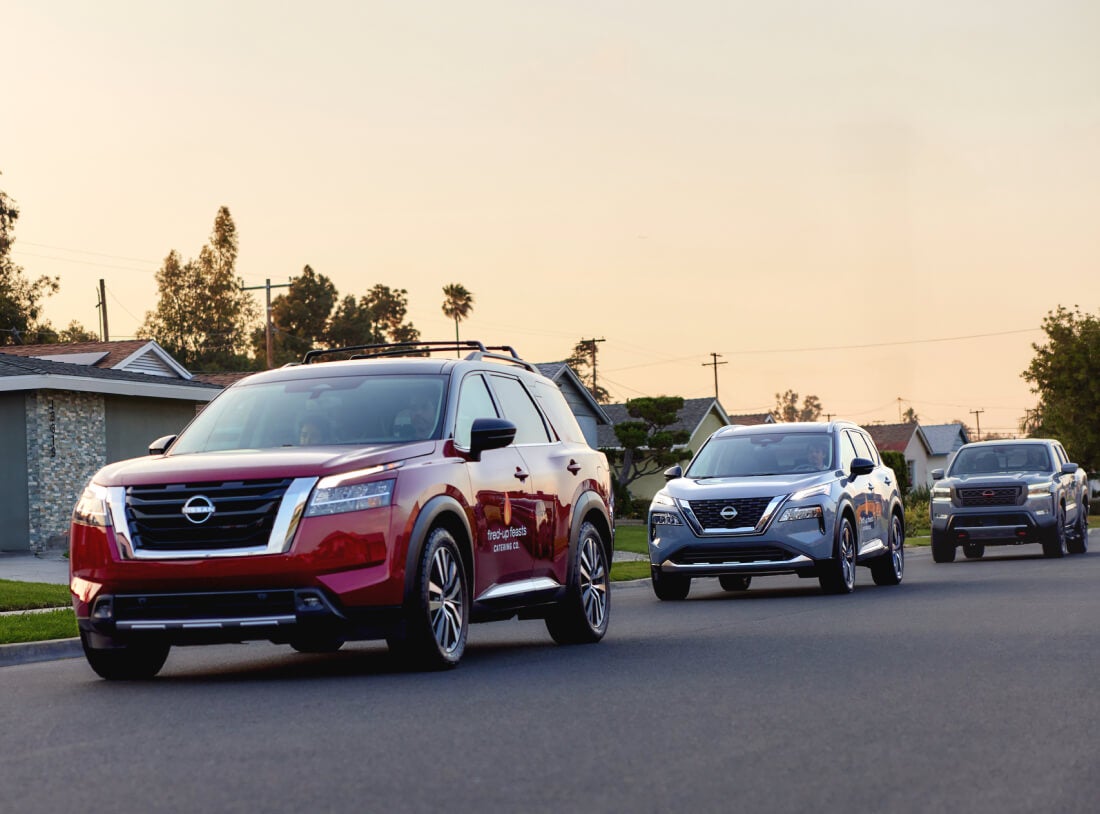 Front three-quarter view of red Pathfinder, silver Rogue, and grey Frontier driving on a suburban road at sunset.